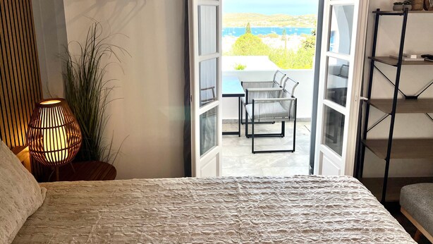Une chambre à coucher avec un lit, une lampe de chevet et une vue sur un balcon extérieur équipé d’une table et de chaises.
