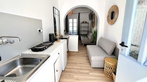 Une cuisine compacte équipée d’un évier, d’une cuisinière et de meubles blancs. Un canapé gris et une petite table d’appoint en osier se trouvent dans l’espace salon, avec vue sur la salle à manger.