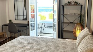 Hotelkamer met een bed, bureau, stoel, boekenkast en uitzicht op de zee door een geopende deur.