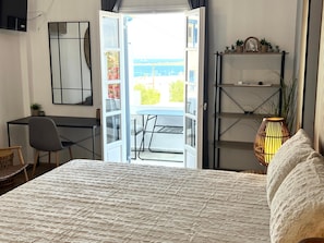 A hotel room with a bed, a desk, a chair, a bookshelf, and a view of the sea through an open door.