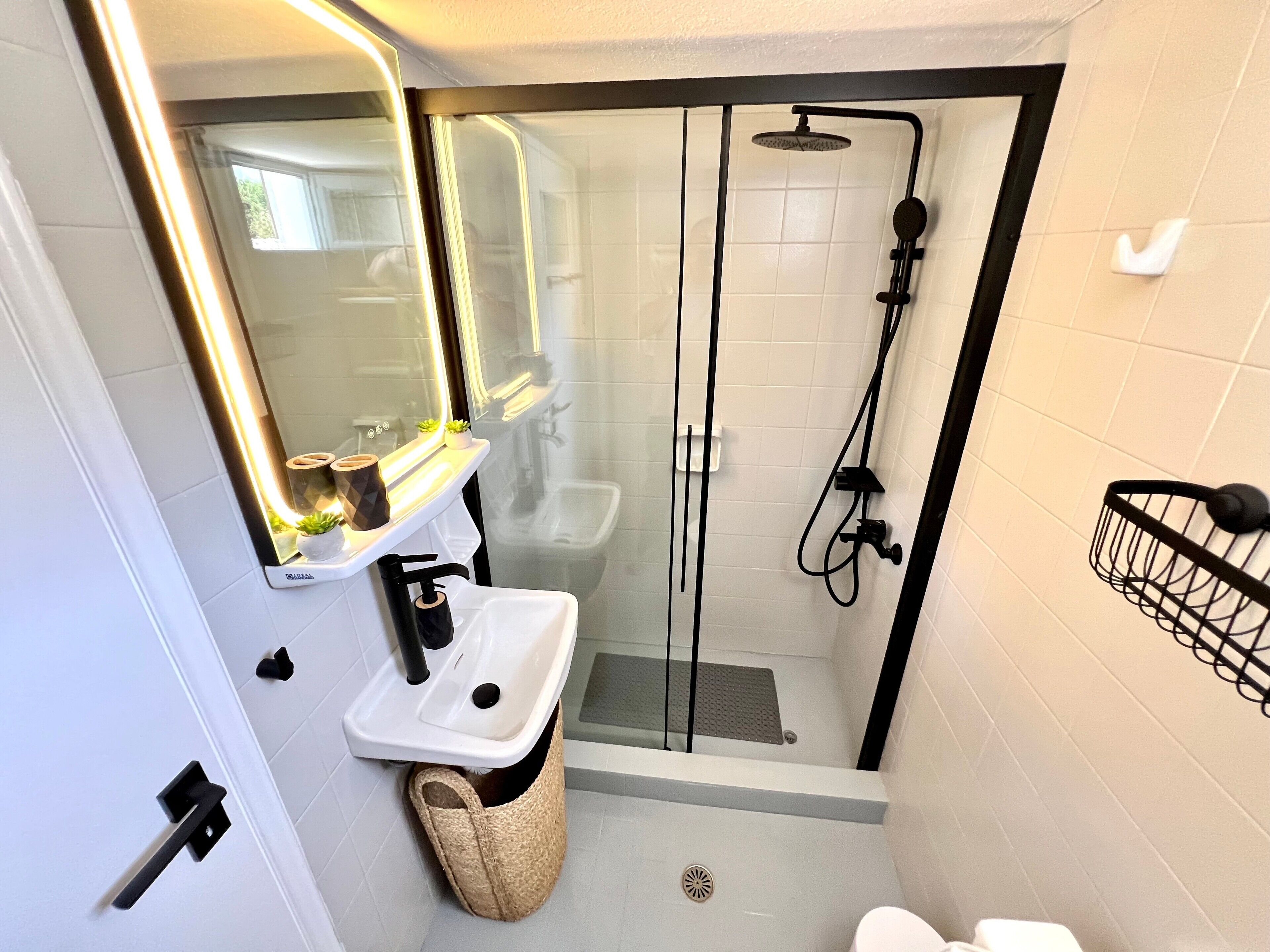 Une salle de bain compacte dotée d’une douche à parois de verre, d’un lavabo blanc et d’un miroir avec éclairage intégré.
