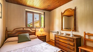 A bedroom with a wooden ceiling, a bed with a green pillow, a dresser, a mirror, and a window with a view of a house.