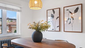 Ein Essbereich mit einem runden Holztisch, vier Korbstühlen, einer großen, dunklen Vase mit Blumen und zwei gerahmten Wandbildern.