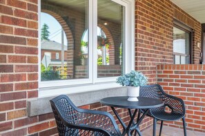 A patio with a brick wall, a window, and two black metal chairs around a small round table with a potted plant. - Upscale Toronto Home Near Transit | Office | King Beds | Garden | Backyard Oasis (Toronto)