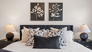 Ein ordentlich bezogenes Bett mit dunkelbrauner Kopfteil, begleitet von zwei Nachttischen mit Lampen, und zwei an der Wand hängenden Kunstwerken mit floralen Motiven.