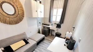 En kompakt opholdsstue med en grå sofa, et lille hvidt bord, et tekøkken med vask og bordplade samt et vindue med gardiner.