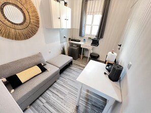 Un espace de vie compact comprenant un canapé gris, une petite table blanche, un coin cuisine avec un évier et un plan de travail, et une fenêtre avec des rideaux.