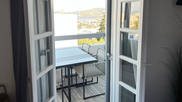 En åben balkon med et bord og stole, udsigt over vand, og en bygning i det fjerne.