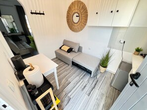 A compact living space with a grey sofa, a small table with a lamp, a coffee machine, and a potted plant.