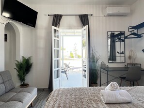 A hotel room with a bed, a desk, a chair, a TV, a balcony with a view, and a potted plant.