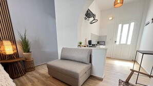 A modern living room with a grey sofa, a small side table, a woven stool, a woven chair, a potted plant, and a kitchen area in the background.