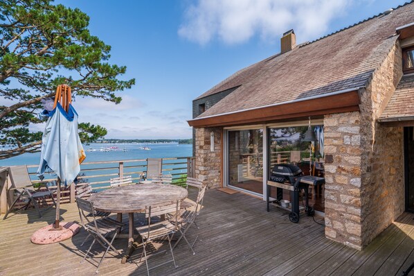 A wooden deck with a table and chairs, a grill, and a view of the water.