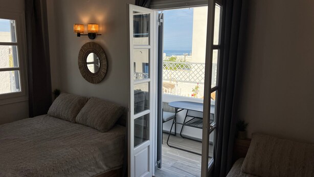 A bedroom with a bed, a bedside table, a mirror, and a door leading to a balcony with a table and chairs. - Studio 'Aeolian Blu 11' with Sea View, Wi-Fi and Air Conditioning (Paros)