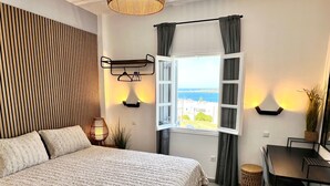 A bedroom with a bed, a desk, a chair, a window with curtains, and a view of the sea.