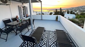 A terrace with a table and chairs, a view of the sunset, and a white building in the background.