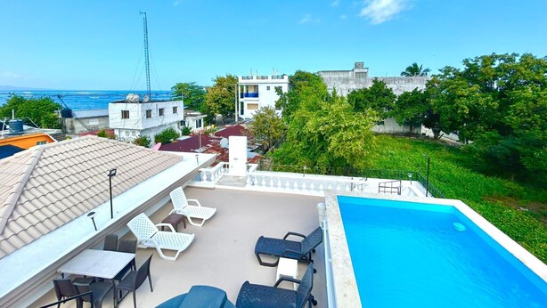 Zona de piscina en la azotea con sillones de descanso y una mesa, con vista a una playa y edificios.