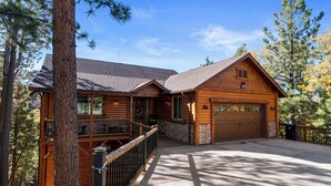 A log cabin with a stone foundation, a large garage, and a deck overlooking a forested area. - Shem Shack (Big Bear City)