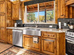 A kitchen with wooden cabinets, a stainless steel sink, a dishwasher, a stove, and a window with a view of trees. - Shem Shack (Big Bear City)