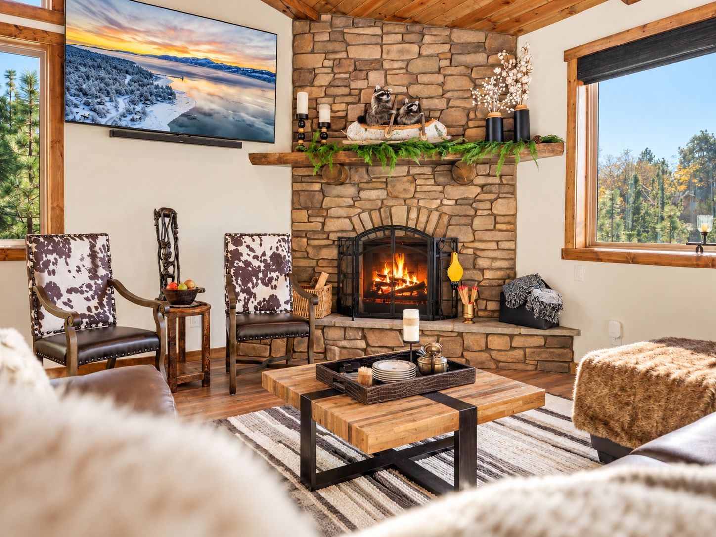 A cozy living room with a stone fireplace, two armchairs, a coffee table, and a large TV mounted on the wall.