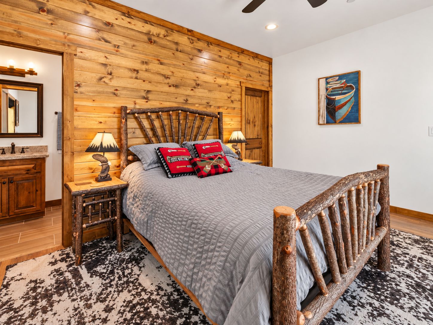 Un dormitorio con cabecera de madera, cama con colcha gris, dos lámparas de noche, mesita de noche de madera, un espejo y un cuadro en la pared.
