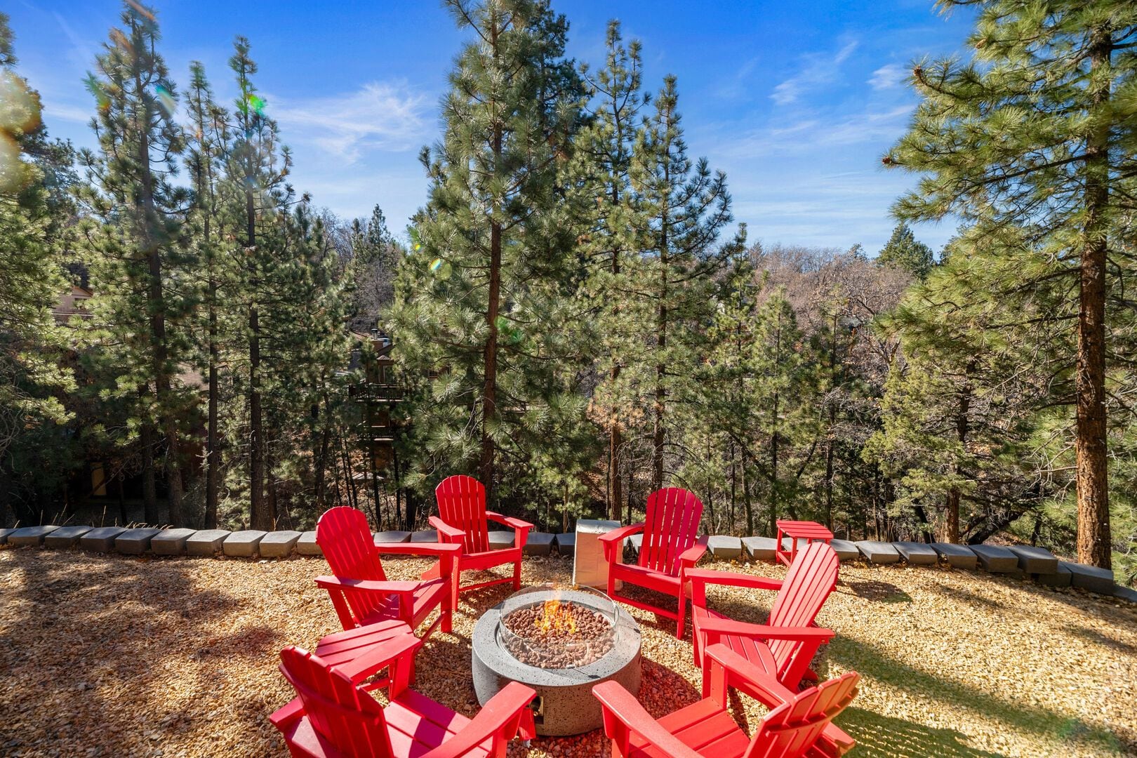 A circular outdoor seating area with red chairs around a central fire pit, surrounded by a forested landscape.