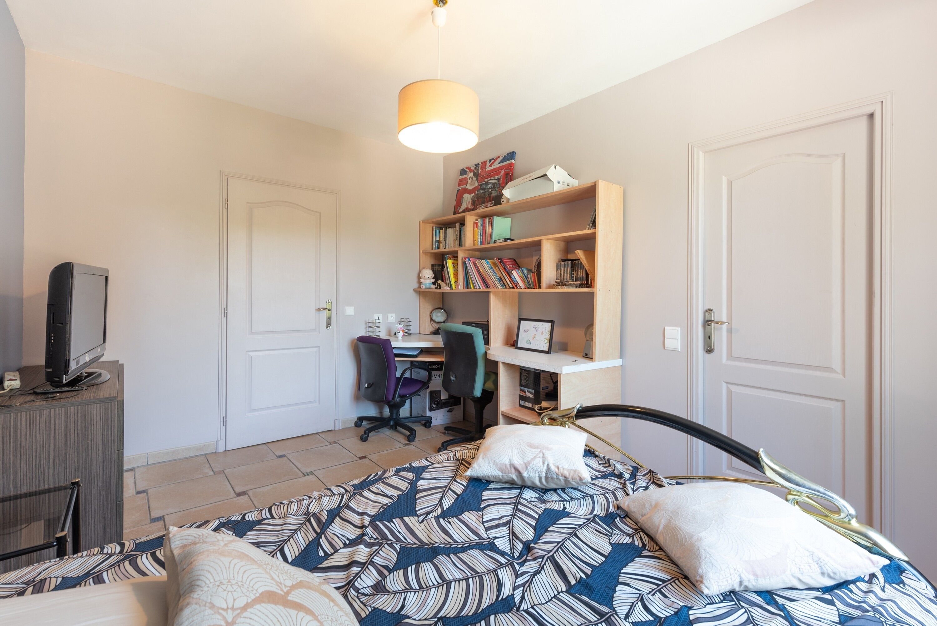 Una camera da letto con un letto, una scrivania con un computer, una sedia, una libreria e una televisione su un mobile.