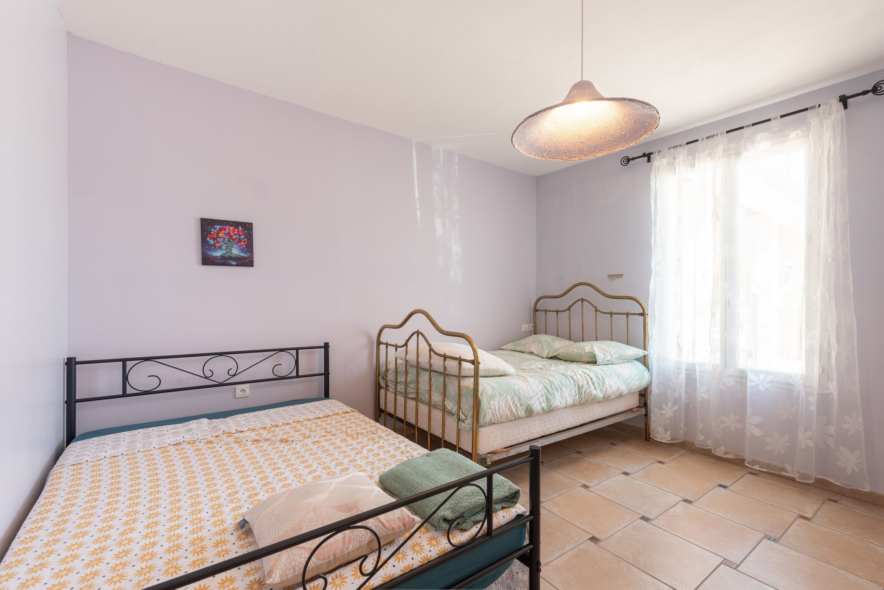 A bedroom with two beds, a hanging light fixture, a window with sheer curtains, and a wall-mounted picture.