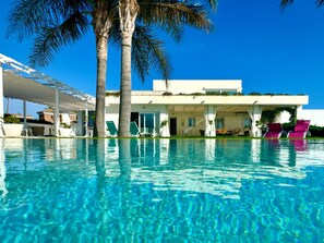 A swimming pool with clear water, palm trees, and a white building with a covered patio area.