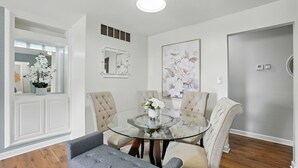 Een eethoek met een glazen tafel, omringd door gestoffeerde stoelen, een potorkidee en een lijstwerk met bloemkunst aan de muur.