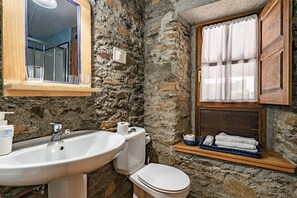 A bathroom with a stone wall, a sink, a toilet, a mirror, and a window with a curtain. - Rural House 'Casa Mario' with Mountain Views, Garden and Wi-Fi (Cangas del Narcea)