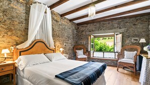 Quarto com parede de pedra, teto de madeira, cama com roupa de cama branca, manta azul e cadeiras de vime.