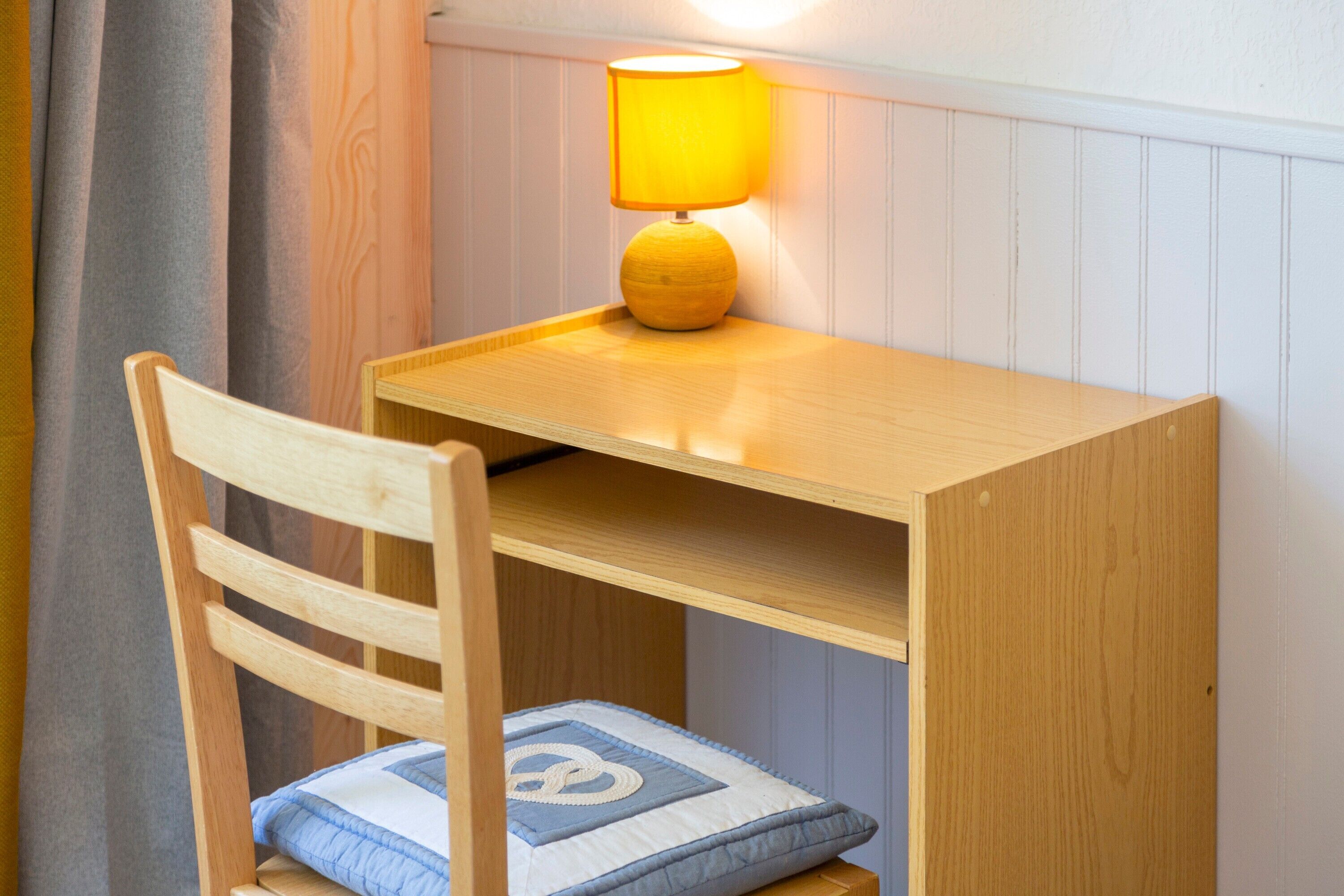 A wooden desk with a lamp, a chair with a cushion, and a wall with paneling.