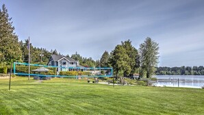 A well-maintained grassy area with a blue volleyball net, a house in the background, and a body of water. - Good Life (lower Suite) @ Shawnigan Beach Resort. (Shawnigan Lake)