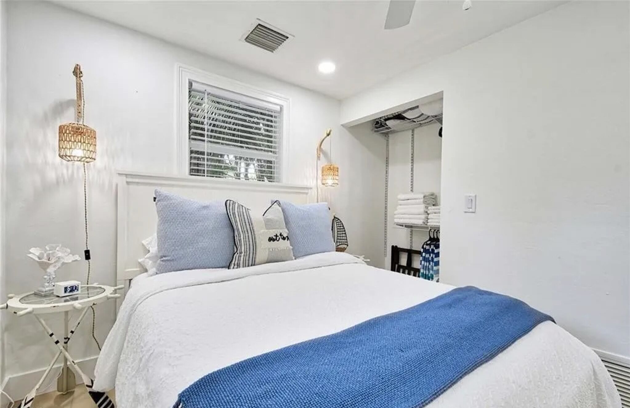 A bedroom with a bed, bedside tables, a window with blinds, a mirror, and a towel rack.