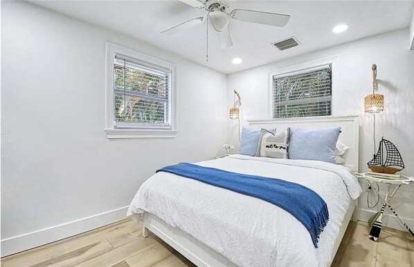 A bedroom with a bed, two windows, a ceiling fan, and a nightstand with a lamp.