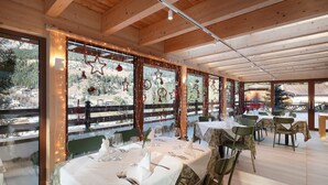 Un espace repas avec des tables préparées pour deux personnes, des poutres en bois au plafond et de grandes fenêtres ornées de luminaires décoratifs.