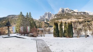 Un sentier enneigé mène à un bâtiment doté d’une terrasse en bois, entouré d’arbres et avec des montagnes en arrière-plan.