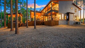 Une maison moderne à deux étages, dotée d’une terrasse en bois et de plusieurs balcons, entourée de grands arbres.