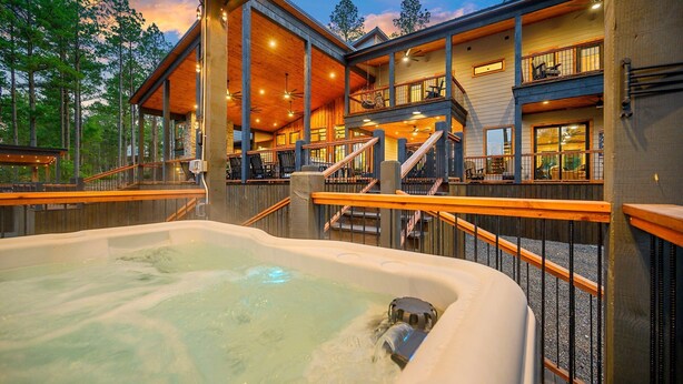 Un espace avec un bain à remous offrant une vue sur un bâtiment de style chalet en bois, des sièges extérieurs et un escalier menant à un balcon.