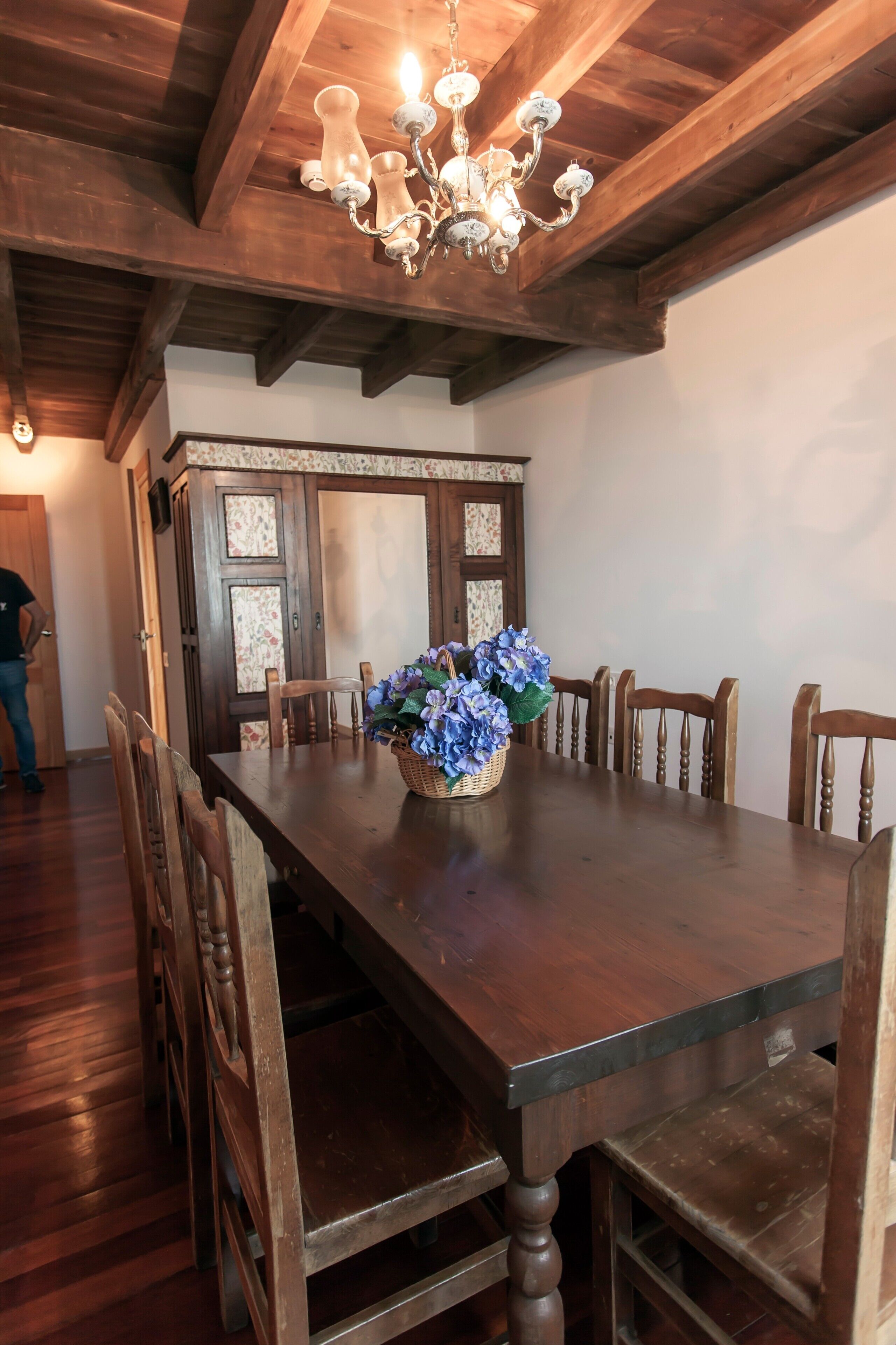 A dining area with a wooden table, chairs, a chandelier, and a floral centerpiece.