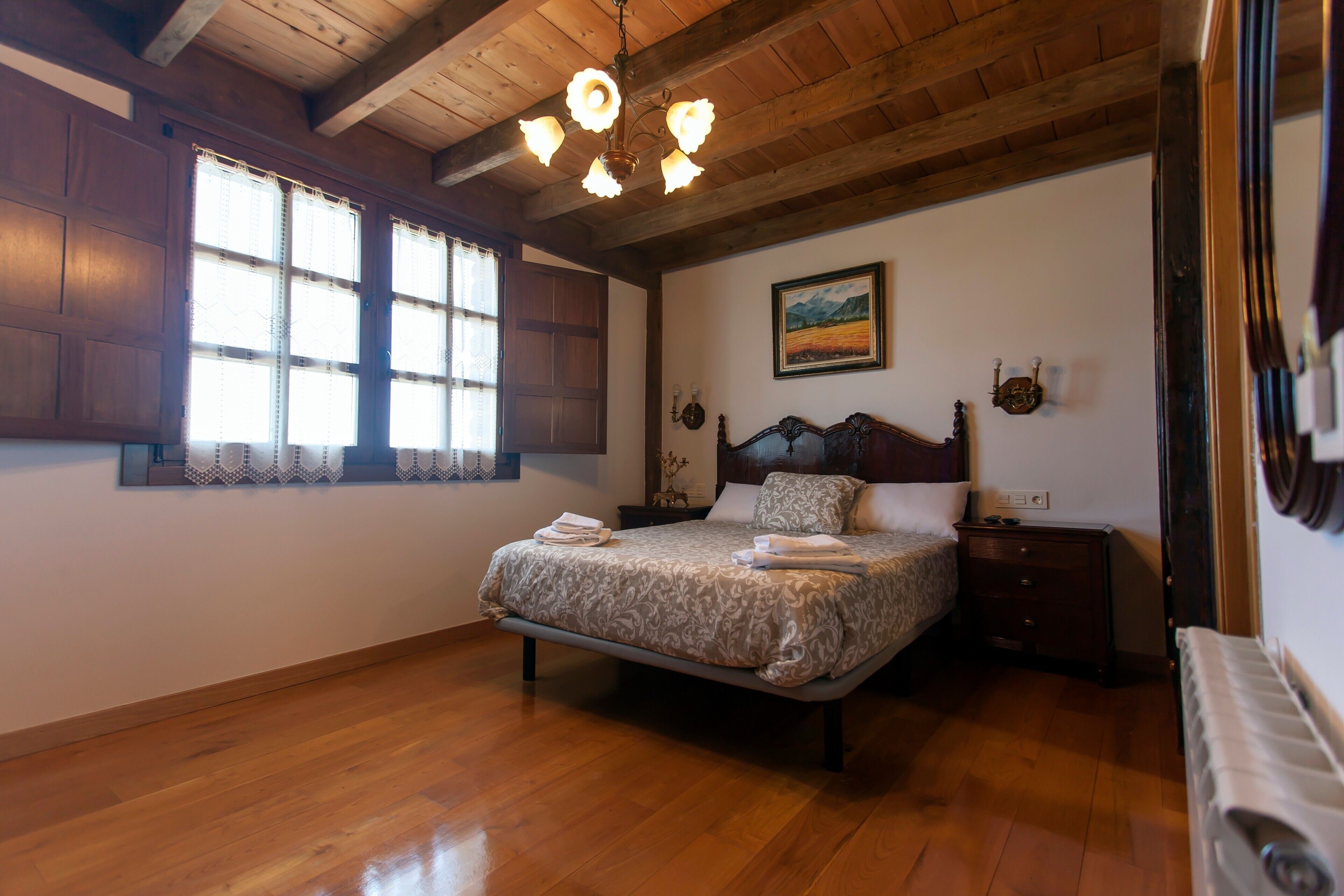 A bedroom with a wooden ceiling, a bed with a patterned bedspread, a nightstand, a wall-mounted lamp, and a painting.