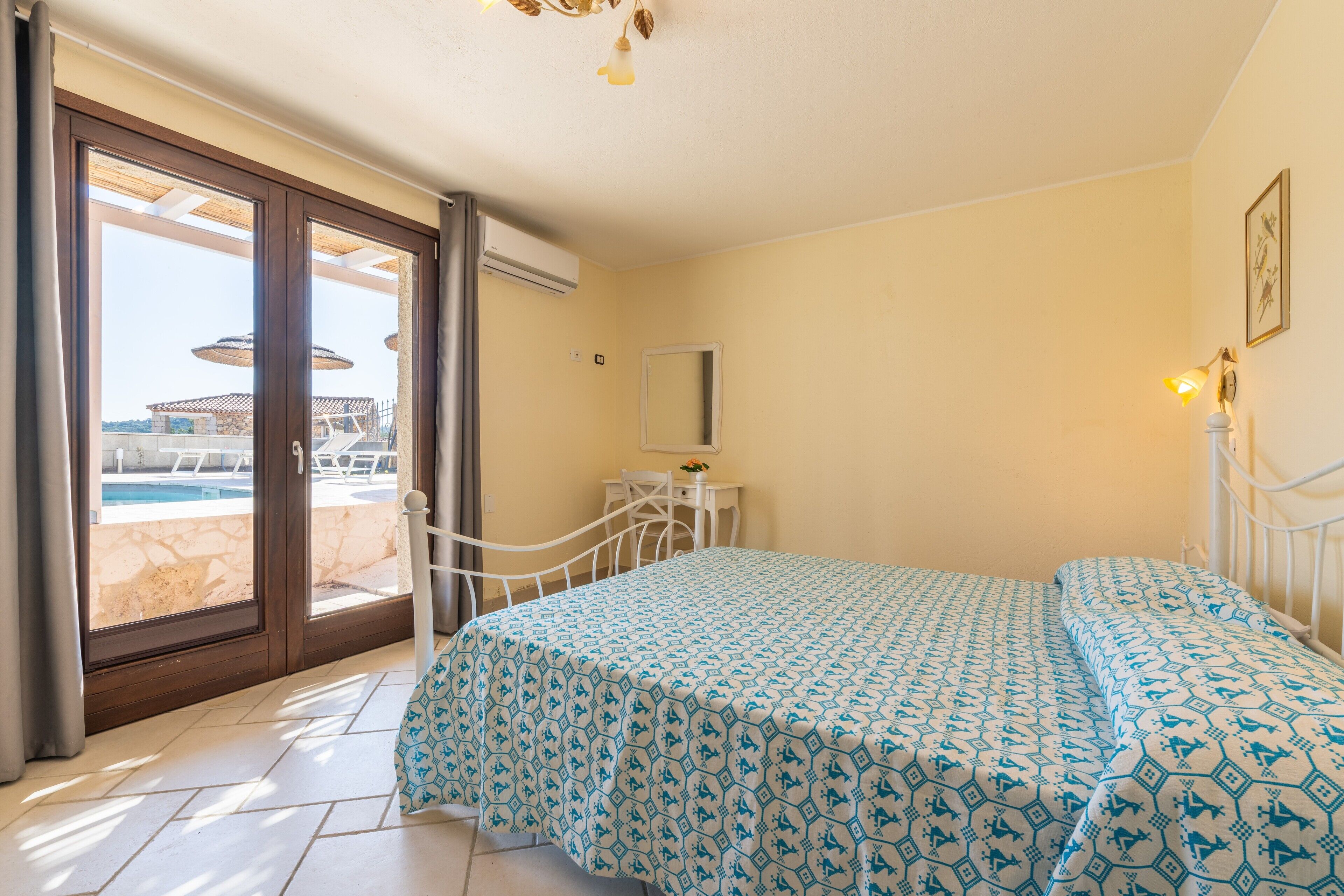 Un dormitorio con cama, escritorio, silla, ventana con vista a una piscina y zona de estar exterior, y ventilador de techo.