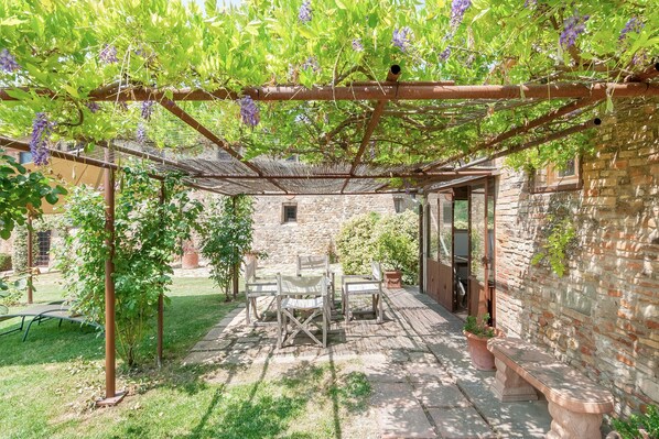 Une terrasse couverte avec une table et des chaises, un mur en pierre et une pergola ornée de fleurs violettes.
