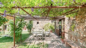 En täckt uteplats med ett bord och stolar, en stenmur och en pergola som är prydd med lila blommor.