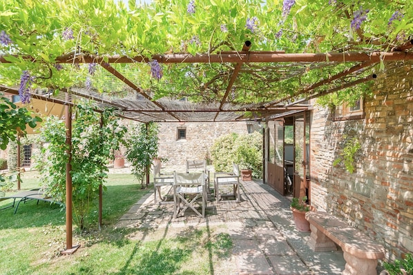 Une terrasse couverte avec une table et des chaises, un mur en pierre et une pergola ornée de fleurs violettes.