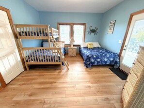 A room with a bunk bed, a single bed, a window with blinds, a bedside table, and a wall decoration.