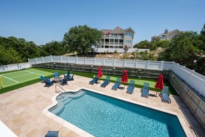 A swimming pool with lounge chairs and umbrellas, a tennis court, and a large house in the background.