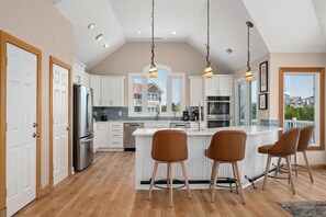 A modern kitchen with white cabinets, stainless steel appliances, a central island with bar stools, and a view of a residential area through large windows. - A Shore Thing: Private Pool, Hot Tub, Elevator, Currituck Club Amenities (Corolla)