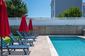 Une piscine entourée de chaises longues et de parasols rouges, le tout délimité par une clôture en bois, avec un bâtiment visible au loin.