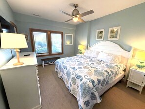 Une chambre à coucher comprenant un lit, des tables de chevet, un bureau, un ventilateur de plafond et une fenêtre avec des stores.